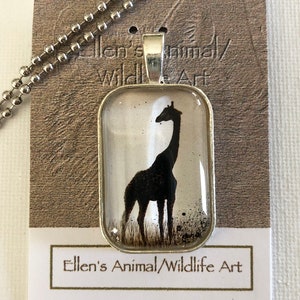 Könnte beinhalten: Eine silberne Kettenkette mit einem rechteckigen Anhänger, der eine schwarze Silhouette einer Giraffe vor weißem Hintergrund zeigt. Der Anhänger ist aus Glas und hat einen silbernen Rahmen. Der Text "Ellen's Animal/Wildlife Art" ist auf der Karte hinter der Kette gedruckt.