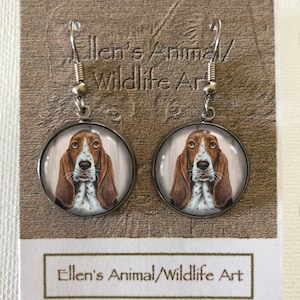 Puede incluir: Un par de pendientes colgantes de plata con un cabujón redondo de vidrio con una imagen pintada de un basset hound marrón y blanco. Los pendientes están en una tarjeta que dice "Ellen's Animal/Wildlife Art".