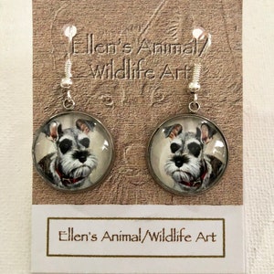 Op de afbeelding: Een paar zilveren oorhangers met een ronde glazen hanger met een geschilderd portret van een dwergkees. De oorbellen bevinden zich op een kaart met de tekst "Ellen's Animal/Wildlife Art".