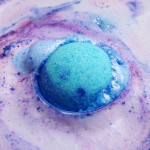 Könnte beinhalten: Nahaufnahme einer blauen Badekugel, die sich in Wasser auflöst und einen schaumigen, farbenfrohen Wirbel erzeugt. Die Badekugel hat eine leuchtende türkise Farbe, umgeben von einer Mischung aus lila und weißen Blasen, die eine Spa-Atmosphäre schaffen.