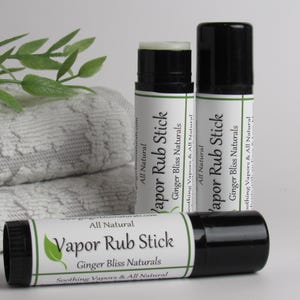 Puede incluir: Tres barras de frotación de vapor con tapas negras y etiquetas blancas, con el texto "Vapor Rub Stick" y "Ginger Bliss Naturals". Un gráfico de hoja verde está en la etiqueta. Una toalla blanca doblada y hojas verdes están en el fondo.