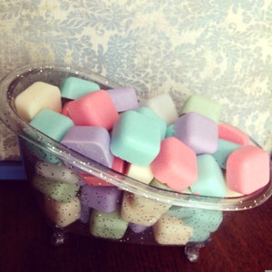 May include: A clear, glitter-edged bathtub filled with pastel-colored, cube-shaped bath bombs. The bath bombs are in shades of pink, blue, purple, and cream. The background features a blue and white floral wallpaper.