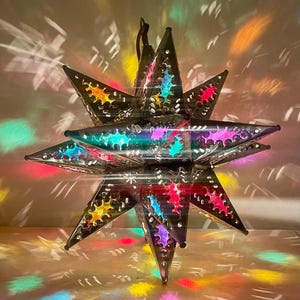 May include: A multi-pointed star-shaped lantern constructed from dark metal panels with intricate cutouts. The cutouts are filled with translucent colored glass in shades of yellow, blue, pink, and green, casting vibrant light patterns.
