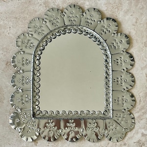 May include: A silver-toned metal mirror featuring an arched glass centre surrounded by a scalloped frame. The frame is decorated with embossed patterns of small dots and wavy lines, creating a textured border around the reflective surface.