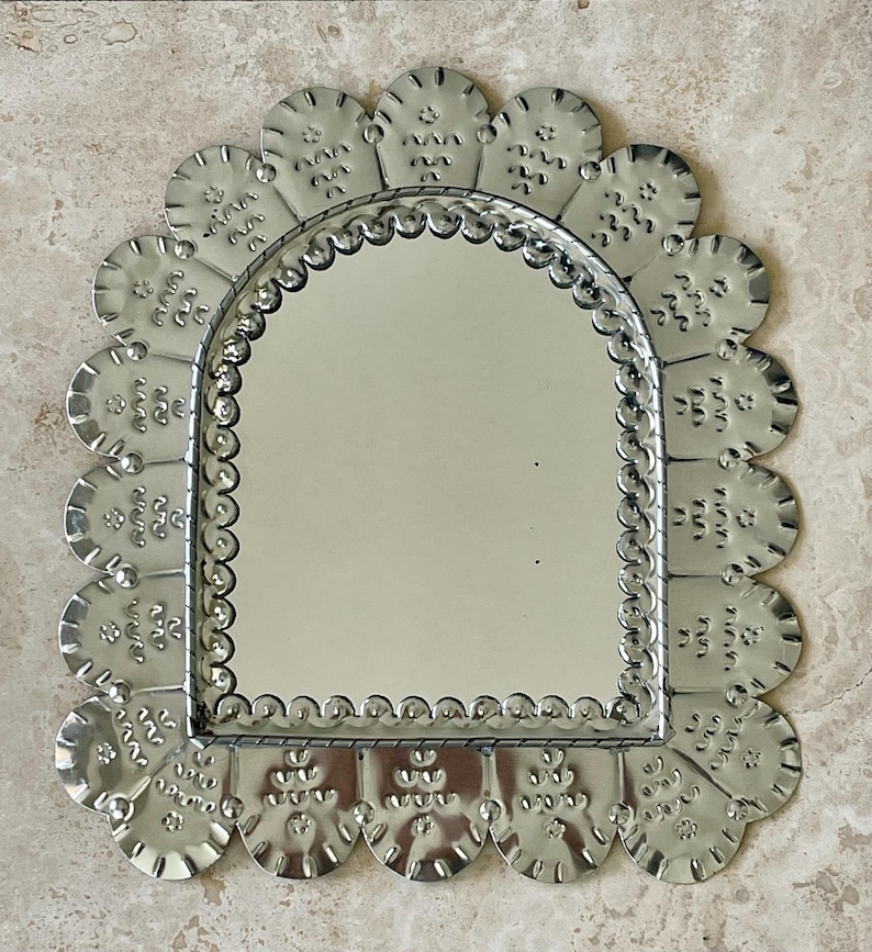 May include: A silver-toned metal mirror featuring an arched glass centre surrounded by a scalloped frame. The frame is decorated with embossed patterns of small dots and wavy lines, creating a textured border around the reflective surface.