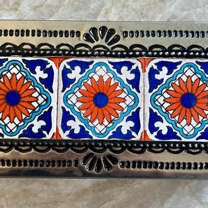 May include: A rectangular metal box with a decorative border and three ceramic tiles. The tiles feature a floral design with orange, blue, and white colors. The metal border has a scalloped edge.