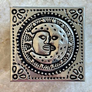 Peut inclure: Boîte carrée en métal argenté avec un motif soleil et lune. Le soleil et la lune sont au centre, entourés d'un cercle noir et d'une bordure décorative. Des motifs floraux ornent les coins.