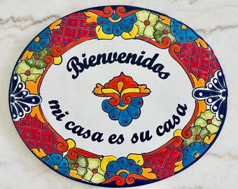 May include: A white oval ceramic plate with a colorful floral border. The plate has the text "Bienvenidos mi casa es su casa" in black lettering.