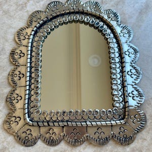 May include: A silver-toned metal wall mirror featuring an arched glass centre. The frame has a scalloped edge with embossed floral-like patterns and a decorative border of small circular indentations surrounding the reflective surface.