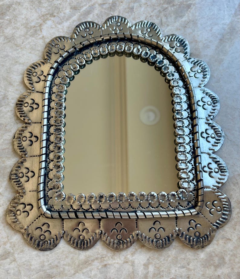 May include: A silver-toned metal wall mirror featuring an arched glass centre. The frame has a scalloped edge with embossed floral-like patterns and a decorative border of small circular indentations surrounding the reflective surface.