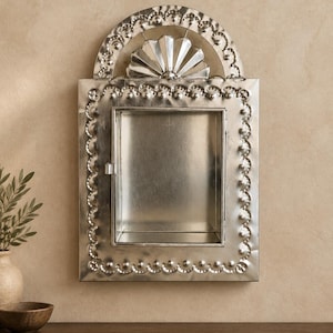 May include: A rectangular silver-toned metal wall cabinet featuring a hinged door with a central recessed compartment. The frame is decorated with a raised beaded border and a semi-circular arch at the top containing a fan-shaped motif.