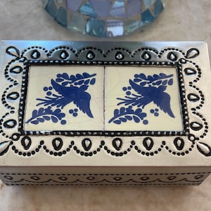 May include: A rectangular silver-toned metal box featuring a decorative border of embossed dots and teardrop shapes. The lid is inlaid with two cream-colored ceramic tiles, each displaying a blue graphic of a bird perched among leafy branches.