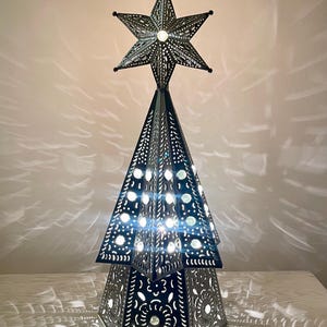 May include: A conical metal lamp shaped like a Christmas tree with intricate cut-out patterns and a star topper. The structure is illuminated from within, casting bright light through the perforations and creating a patterned glow.