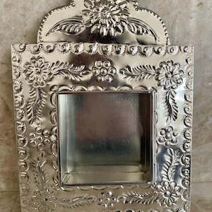 May include: A silver-colored metal wall-mounted display box featuring an arched top and a rectangular central opening. The frame is decorated with embossed floral patterns, leaves, and small circular dots throughout the entire surface.
