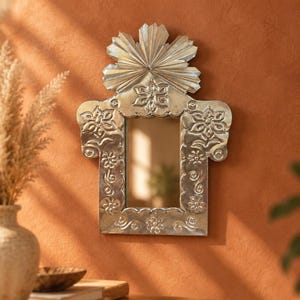 May include: A rectangular wall mirror with a silver-toned metal frame featuring embossed floral and swirling patterns. The top of the frame is adorned with a prominent, fan-shaped crest. The central glass is clear and reflective.