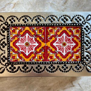 May include: Rectangular metal box with a decorative border and a colorful tile design. The tile features a red and white floral pattern on a yellow background. The border is silver with black accents.