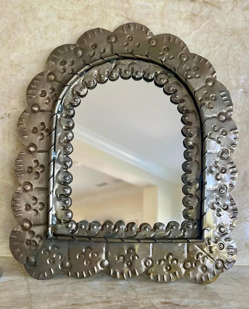 May include: An arched wall mirror featuring a wide, scalloped metal frame with a brushed silver finish. The frame is decorated with embossed floral and circular patterns, and a twisted metal border surrounds the central reflective glass surface.