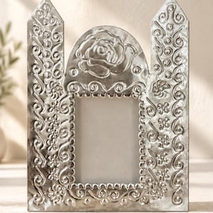 May include: A silver-coloured metal picture frame with a rectangular centre opening. The frame features embossed floral patterns, swirling vine motifs, and a prominent rose design at the top arch, with two pointed towers on the sides.
