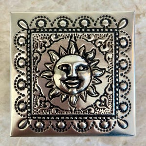 May include: Square, silver-colored metal box with a detailed sun face design in the center. The sun has a smiling face and is surrounded by a decorative border. The box has an ornate, patterned edge.
