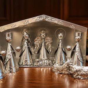 May include: A silver-toned metal nativity set featuring a stable structure, four standing figures with halo accents, and two animal figurines. The figures have conical bases and metallic textures, with two figures holding small circular candle holders.
