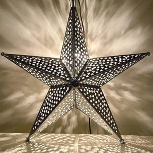 May include: A silver metal star-shaped light fixture with a cut-out design of smaller stars. The light is on and casting a pattern of light on the surface below.