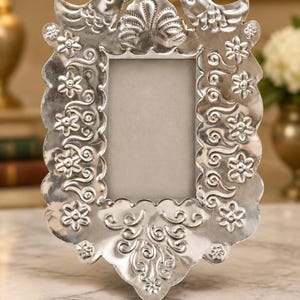 May include: A rectangular silver-toned metal picture frame featuring embossed floral patterns, swirling vine motifs, and two bird figures at the top corners. The frame has a scalloped edge and a central rectangular opening for a photograph.