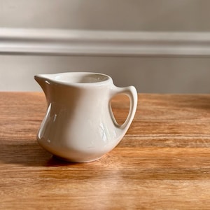 May include: A small, off-white ceramic creamer pitcher with a smooth, glossy finish. The vessel features a rounded body, a short pouring spout, and a single curved handle on the side. The design is simple and functional, with a solid color throughout.