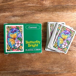 May include: A green deck box labeled Current Butterfly Bright Playing Cards next to a fanned stack of playing cards. Each card features a colorful stained glass style illustration of a yellow and blue butterfly surrounded by pink and yellow flowers.