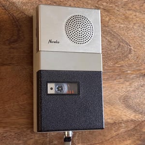 May include: A vintage Norelco portable cassette recorder featuring a two-tone design with a silver top section containing a circular speaker grille and a black textured bottom section with a clear window revealing the internal cassette tape mechanism.