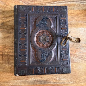 May include: A rectangular dark brown leather-bound book featuring intricate embossed floral and geometric patterns across the cover. The center displays a circular medallion with a star motif, and a tan fabric ribbon is tied to the right side.