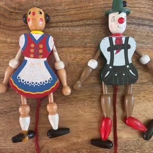 May include: Two wooden jumping jack puppets featuring painted figures in traditional attire. The left figure wears a red dress with a white apron, while the right figure wears a green hat, white shirt, and dark lederhosen. Both have red strings.
