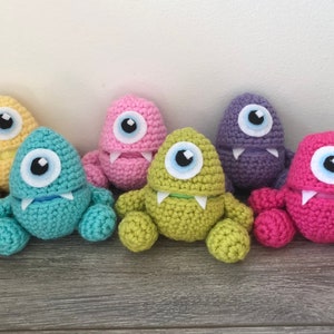 May include: Six colourful, handmade crochet monster toys. Each has a single large eye, a small mouth with teeth, and rounded limbs. The toys are in shades of yellow, teal, pink, green, purple, and magenta. They are displayed on a wooden surface.