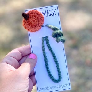 Puede incluir: Un marcapáginas de ganchillo hecho a mano con una calabaza naranja, un tallo marrón y hojas verdes. El marcapáginas tiene un lazo verde y está unido a una tarjeta blanca con la palabra "MARK" y el sitio web "yarnandcountryca.etsy.com".