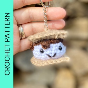 May include: A crocheted s'more keychain with a brown chocolate bar, white marshmallow, and tan graham cracker. The marshmallow has a smiling face with black eyes and a small mouth.