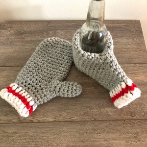 May include: A pair of grey crocheted mittens with white and red striped cuffs. One mitten is laid flat, while the other is upright and holds a clear glass bottle. The yarn texture is visible throughout the grey body and the ribbed cuff design.