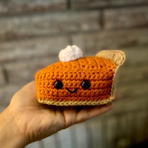 May include: A crocheted slice of pumpkin pie with a smiling face and a dollop of white cream. The pie is orange with a beige crust and a small piece of beige crust on the side. The pie is held in a hand.