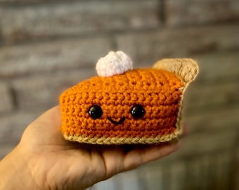 Puede incluir: Una rebanada de tarta de calabaza tejida con una cara sonriente y una bolita de crema blanca. La tarta es naranja con una corteza beige y un pequeño trozo de corteza beige en el lateral. La tarta se sostiene en una mano.