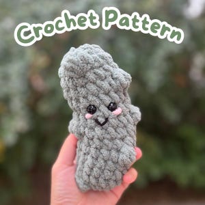 Può includere: Peluche all'uncinetto grigio fatto a mano con un viso sorridente, occhi neri e guance rosa. Il peluche è tenuto in una mano, con la scritta "Crochet Pattern" in una bolla verde sopra. Il peluche ha un aspetto morbido e strutturato.