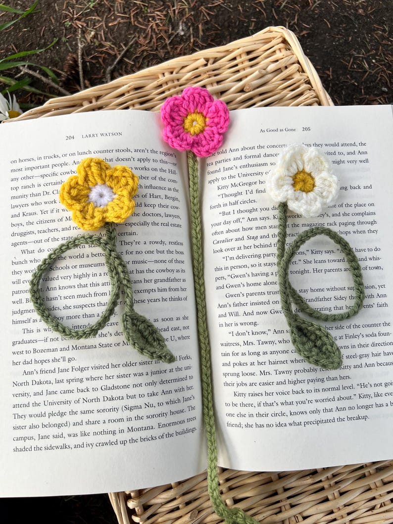 May include: Crocheted flower bookmarks in yellow, pink, and white, resting on an open book. The handmade bookmarks feature detailed flower designs with green stems and leaves, perfect for book lovers.