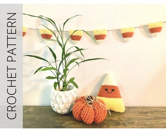 Pode incluir: Uma guirlanda de milho doce de crochê com listras laranja, amarelas e brancas penduradas em uma parede branca. Um travesseiro de milho doce de crochê com um rosto sorridente fica em uma mesa de madeira ao lado de uma abóbora laranja de crochê.