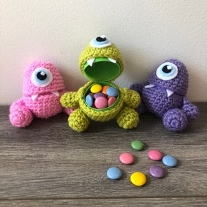 May include: Three crocheted monster-shaped candy holders in pink, green, and purple. The green monster has an open mouth revealing a plastic container filled with colourful sweets. Each monster has a single eye and small teeth. Sweets are scattered around.