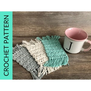 May include: Three crocheted coasters with fringe, in gray, white, and teal, on a wood surface. A pink mug with a white letter J is in the background.