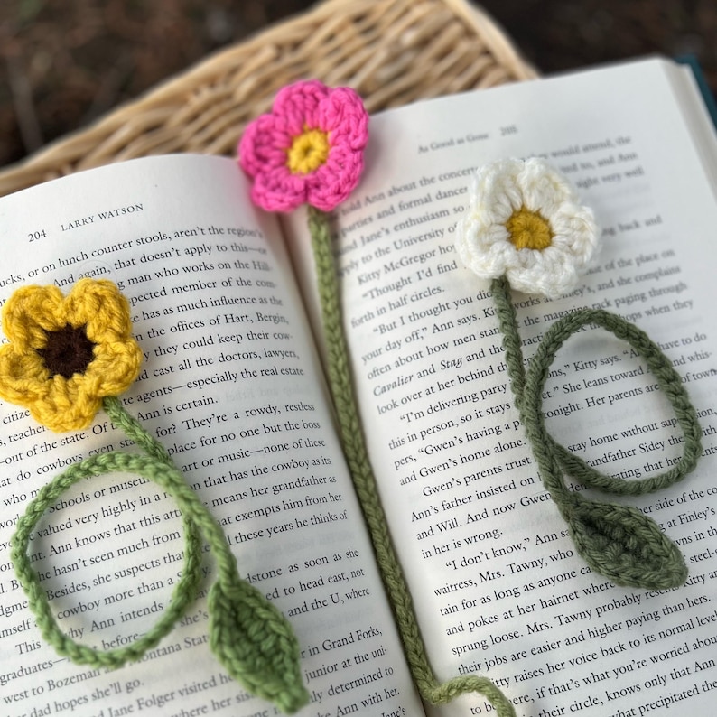 May include: Crocheted flower bookmarks in yellow, pink, and white, with green stems and leaves, resting on the pages of a book. Handmade floral design for book lovers. Each bookmark is approximately 10 cm in length.