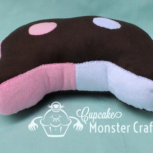 Puede incluir: Una almohada de felpa con forma de orca, con la parte superior marr&oacute;n, manchas rosas y celeste, y laterales rosas y blancos. La almohada tiene una forma curva y est&aacute; dise&ntilde;ada para parecerse a una ballena de dibujos animados. El texto "Cupcake Monster Crafts" es visible.