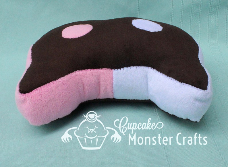 Puede incluir: Una almohada de felpa con forma de orca, con la parte superior marr&oacute;n, manchas rosas y celeste, y laterales rosas y blancos. La almohada tiene una forma curva y est&aacute; dise&ntilde;ada para parecerse a una ballena de dibujos animados. El texto "Cupcake Monster Crafts" es visible.