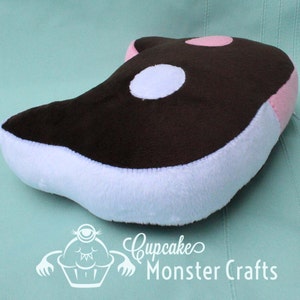 Puede incluir: Un peluche con forma de orca, con la parte superior marr&oacute;n, vientre blanco y detalles rosas. Tiene una mancha blanca y rosa en la parte superior. El juguete est&aacute; dise&ntilde;ado para ni&ntilde;os. El logotipo "Cupcake Monster Crafts" es visible.