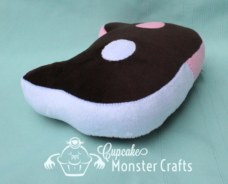Puede incluir: Un peluche con forma de orca, con la parte superior marr&oacute;n, vientre blanco y detalles rosas. Tiene una mancha blanca y rosa en la parte superior. El juguete est&aacute; dise&ntilde;ado para ni&ntilde;os. El logotipo "Cupcake Monster Crafts" es visible.