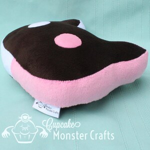 Puede incluir: Un coj&iacute;n de peluche con forma de orca de dibujos animados. El coj&iacute;n presenta una parte superior marr&oacute;n con manchas blancas y rosas, laterales rosas y un acento blanco. La etiqueta dice "Cupcake Monster Crafts".