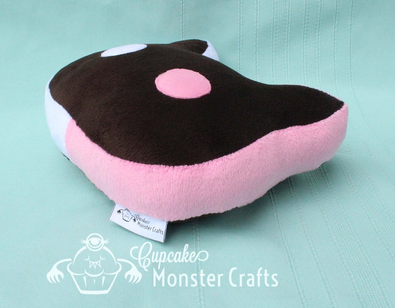 Puede incluir: Un coj&iacute;n de peluche con forma de orca de dibujos animados. El coj&iacute;n presenta una parte superior marr&oacute;n con manchas blancas y rosas, laterales rosas y un acento blanco. La etiqueta dice "Cupcake Monster Crafts".