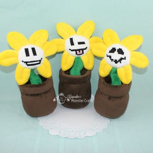 May include: Three plush yellow flower toys with different pixelated faces. Each flower is in a brown pot. The flowers are on a white doily.